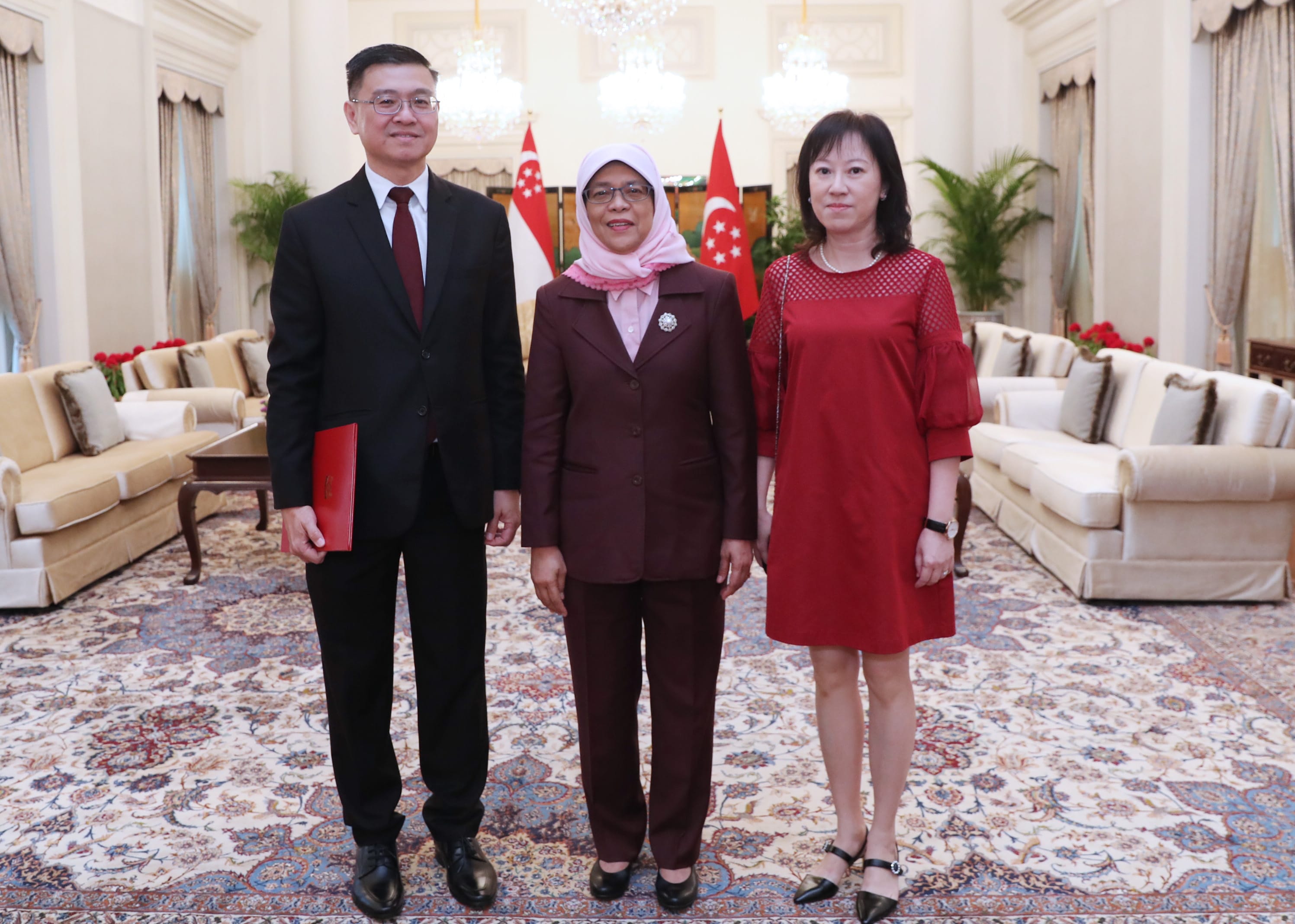 Three people stand before Singapore flags in a room with ornate decor.